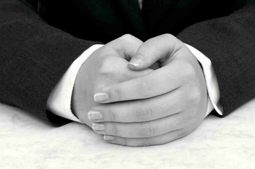 Detail of young woman's hands folded, dark suit and white shirt, hands resting on desk.