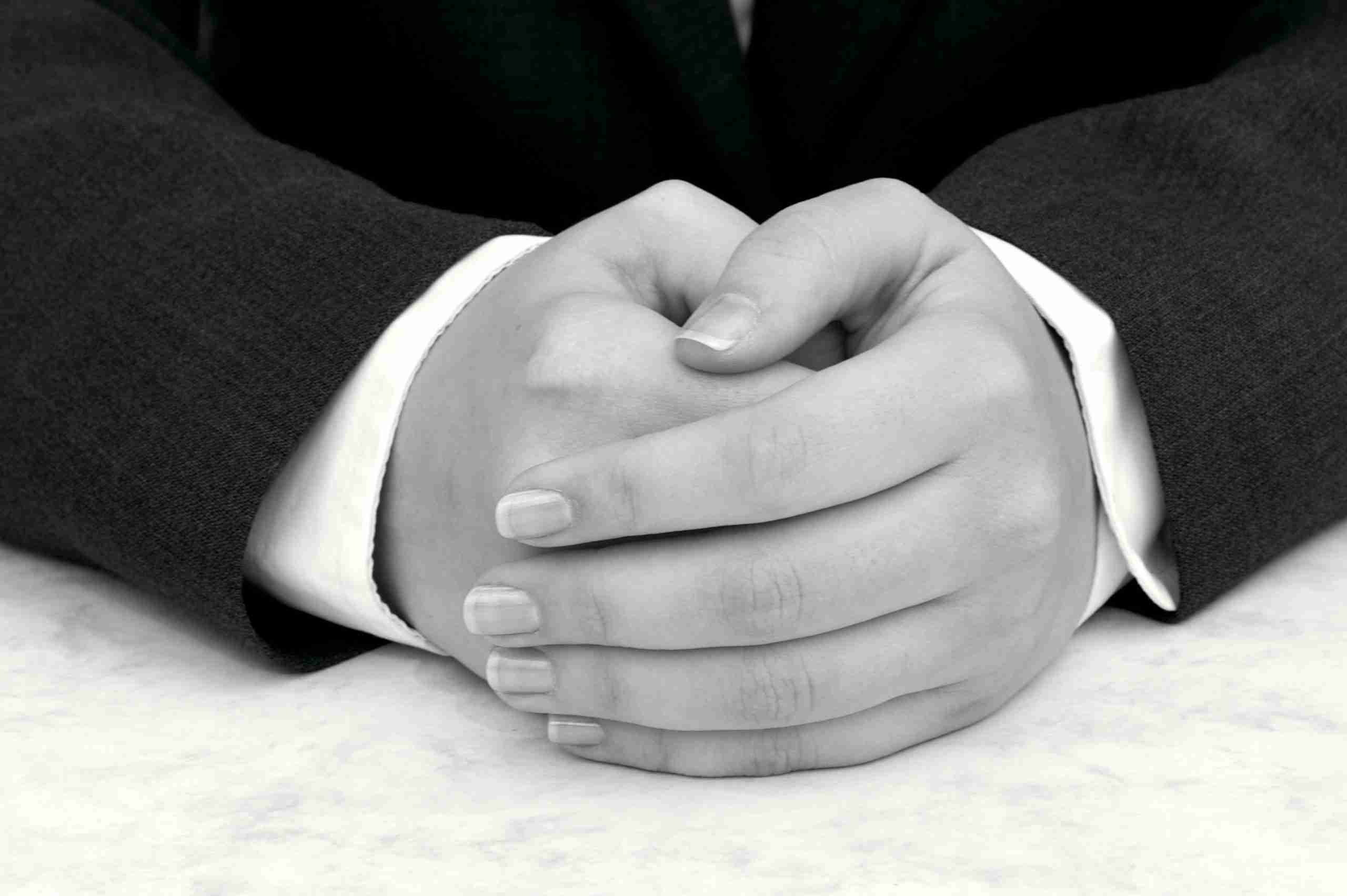Detail of young woman's hands folded, dark suit and white shirt, hands resting on desk.